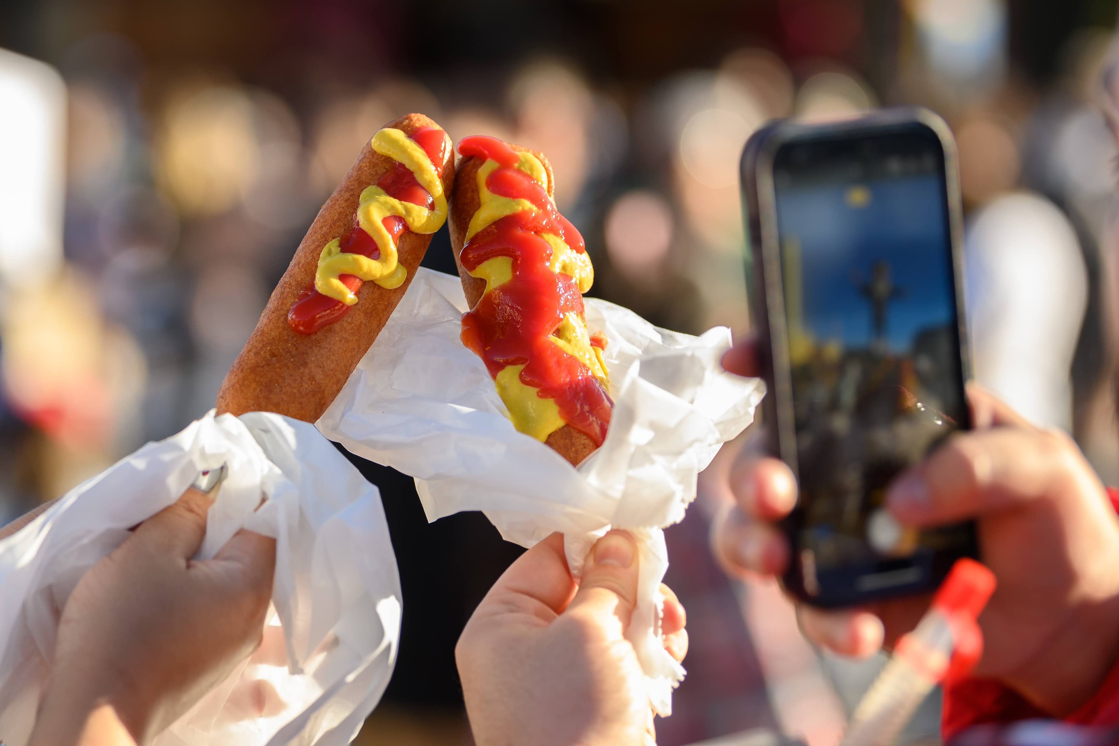 File photo: Fairgoers hold two corny dogs topped with mustard and ketchup; another person photographs them. This archival image does not depict a new 2026 menu item.